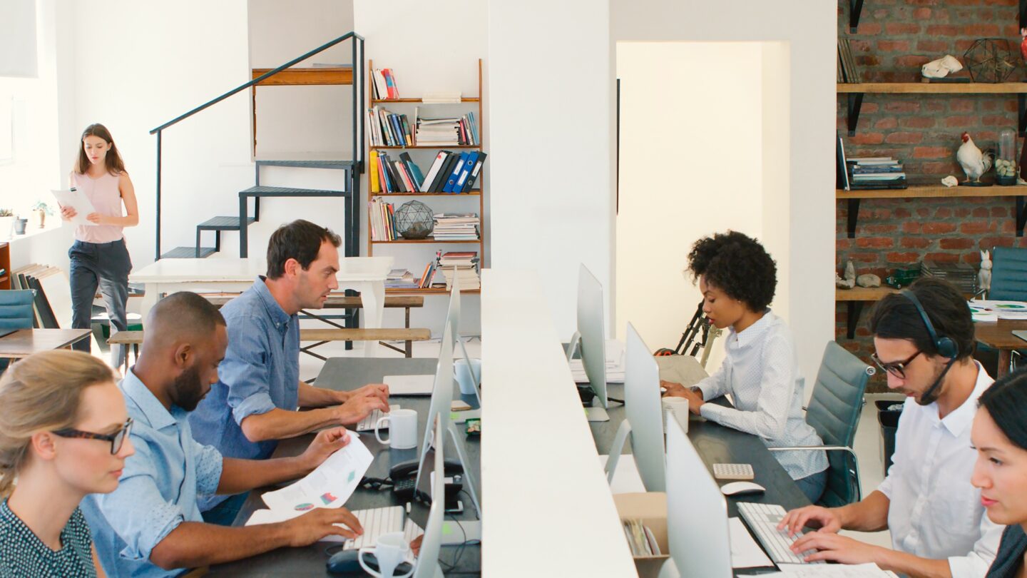 Multi-Cultural Business Team Working At Desks On Computers In Modern Open Plan Office